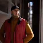 A man wearing a cap stands in a darkened industrial room. He is looking to the side with a serious expression