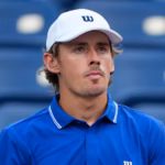 A male tennis player in a blue shirt sits courtside, looking frustrated