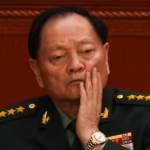 A general in a green jacket holds his hand to his face, with a red wall behind him