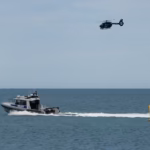 A water police speedboat travels in open ocean near the shore, while a helicopter flies above